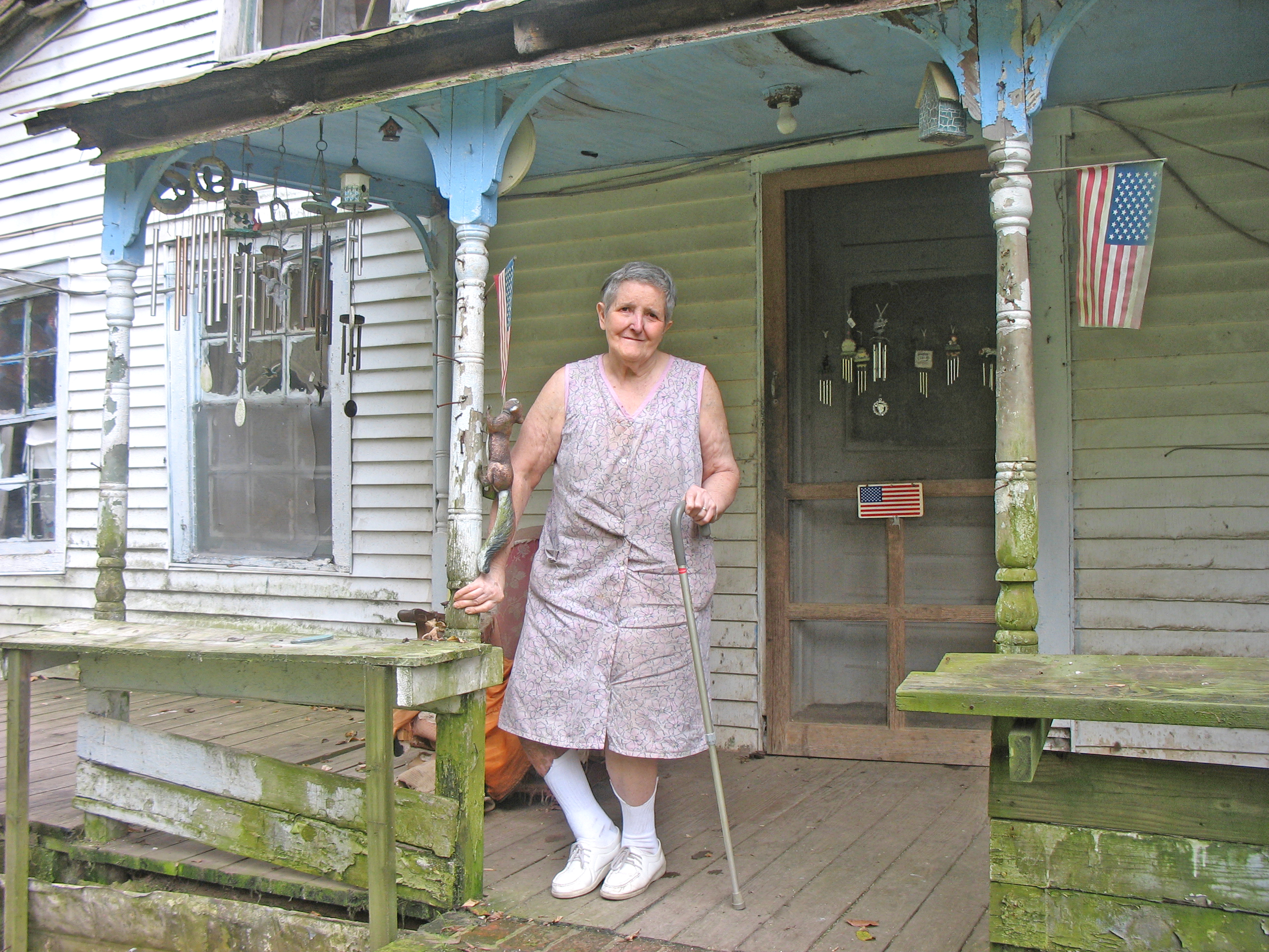 Elderly woman with a cane standing on the front porch of a house