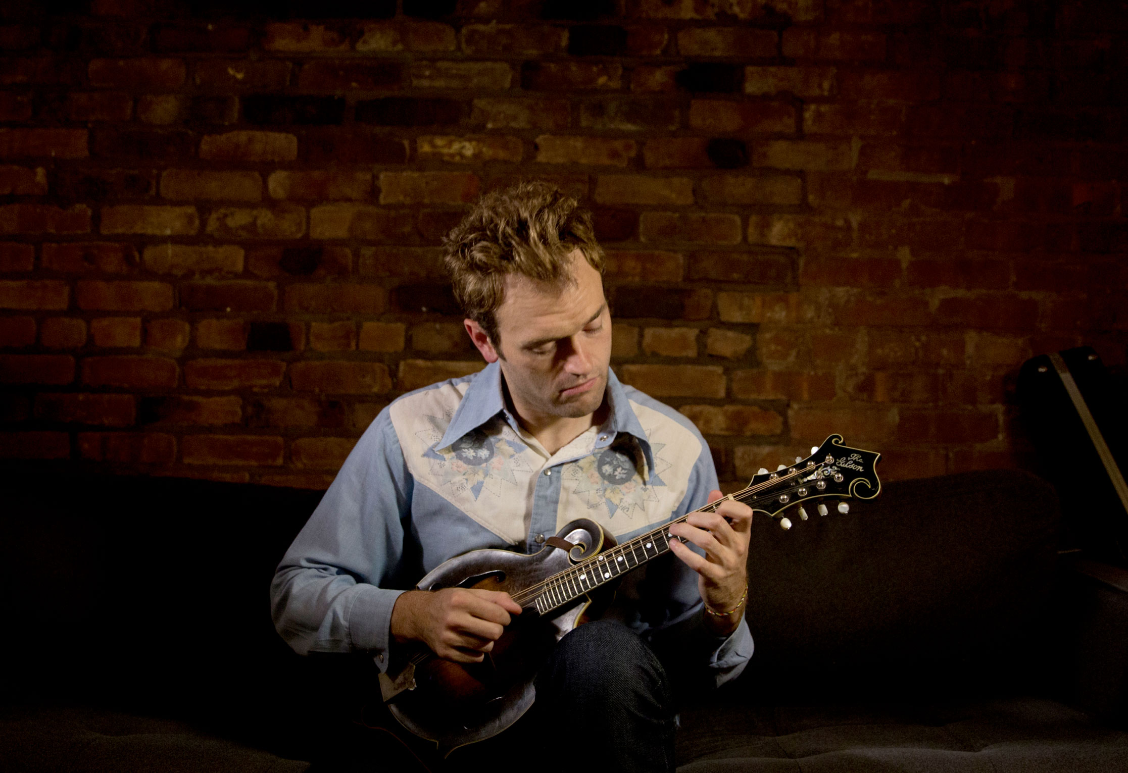 A man with brown hair in a blue and white shirt sits and plays the mandolin.
