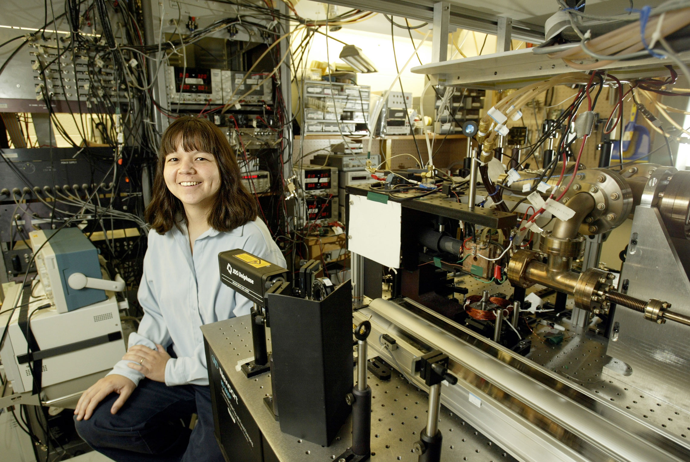 A woman with shoulder length brown hair sits in a Physics lab.