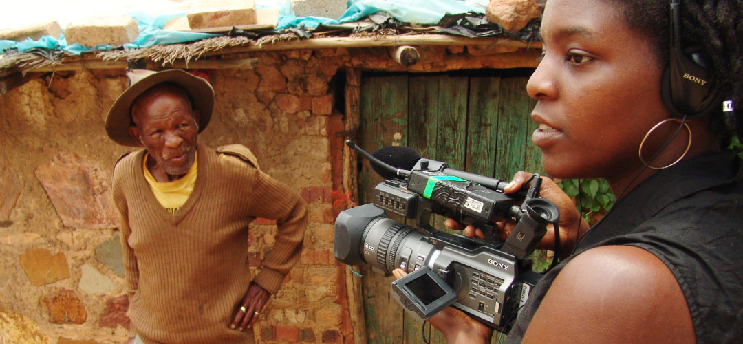A woman holds a camera pointed toward a man during an interview.