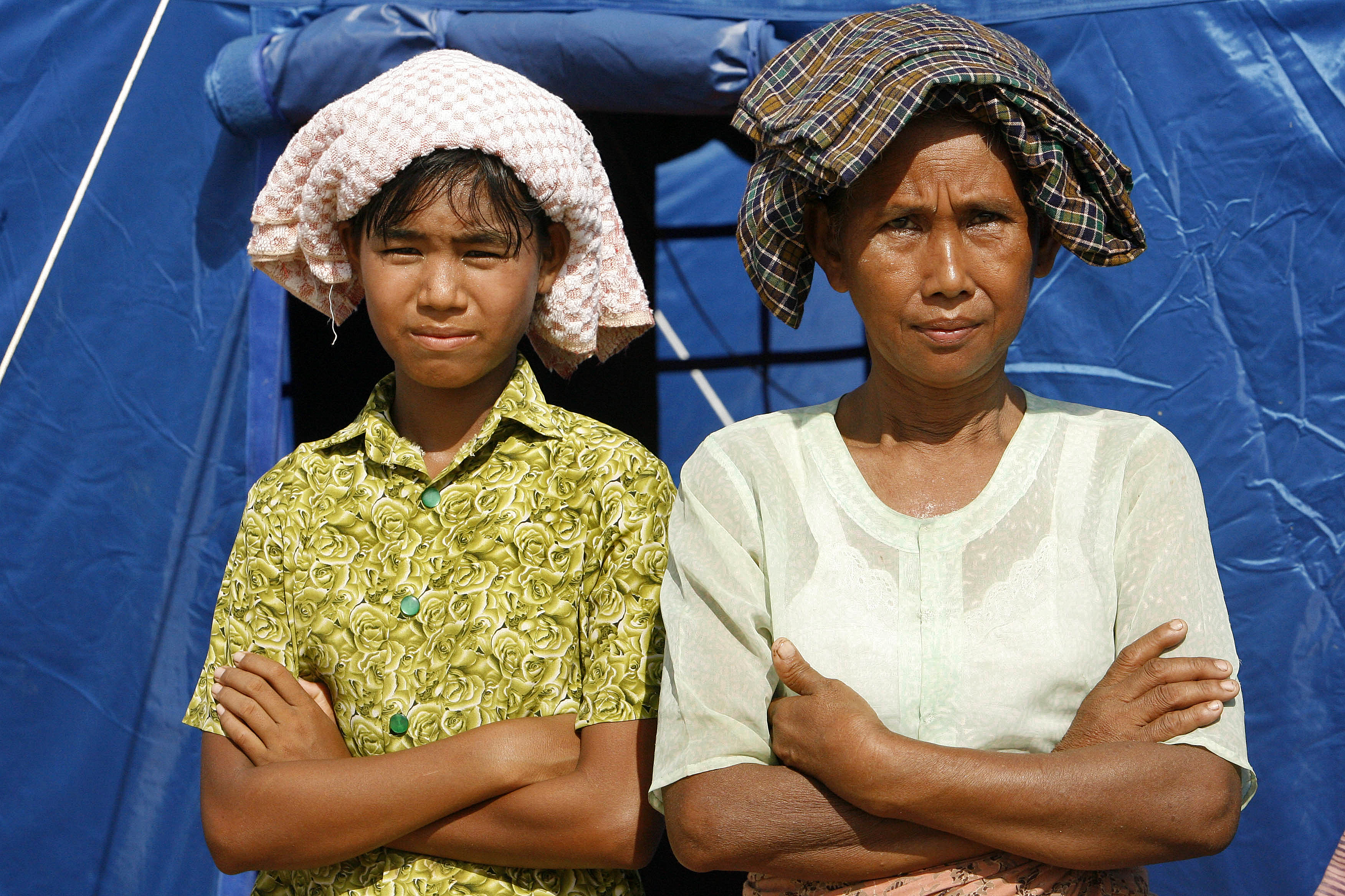 A woman and a young child stand in front of a tent in the sun with cloth on their heads.