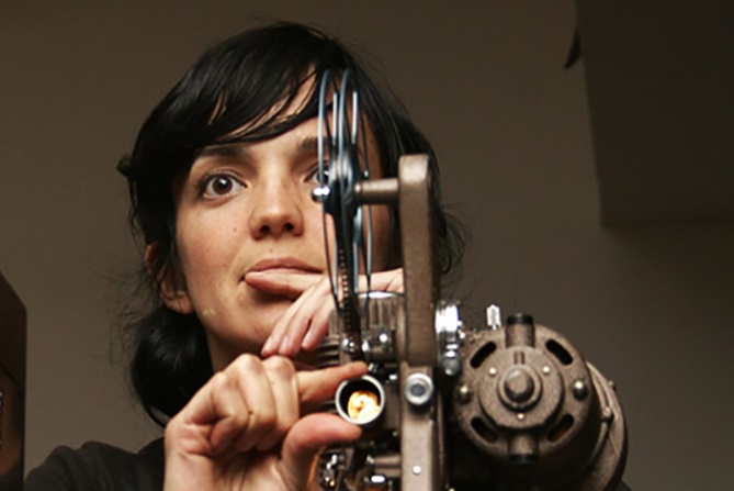 A woman stands behind and holds a film projection tool.