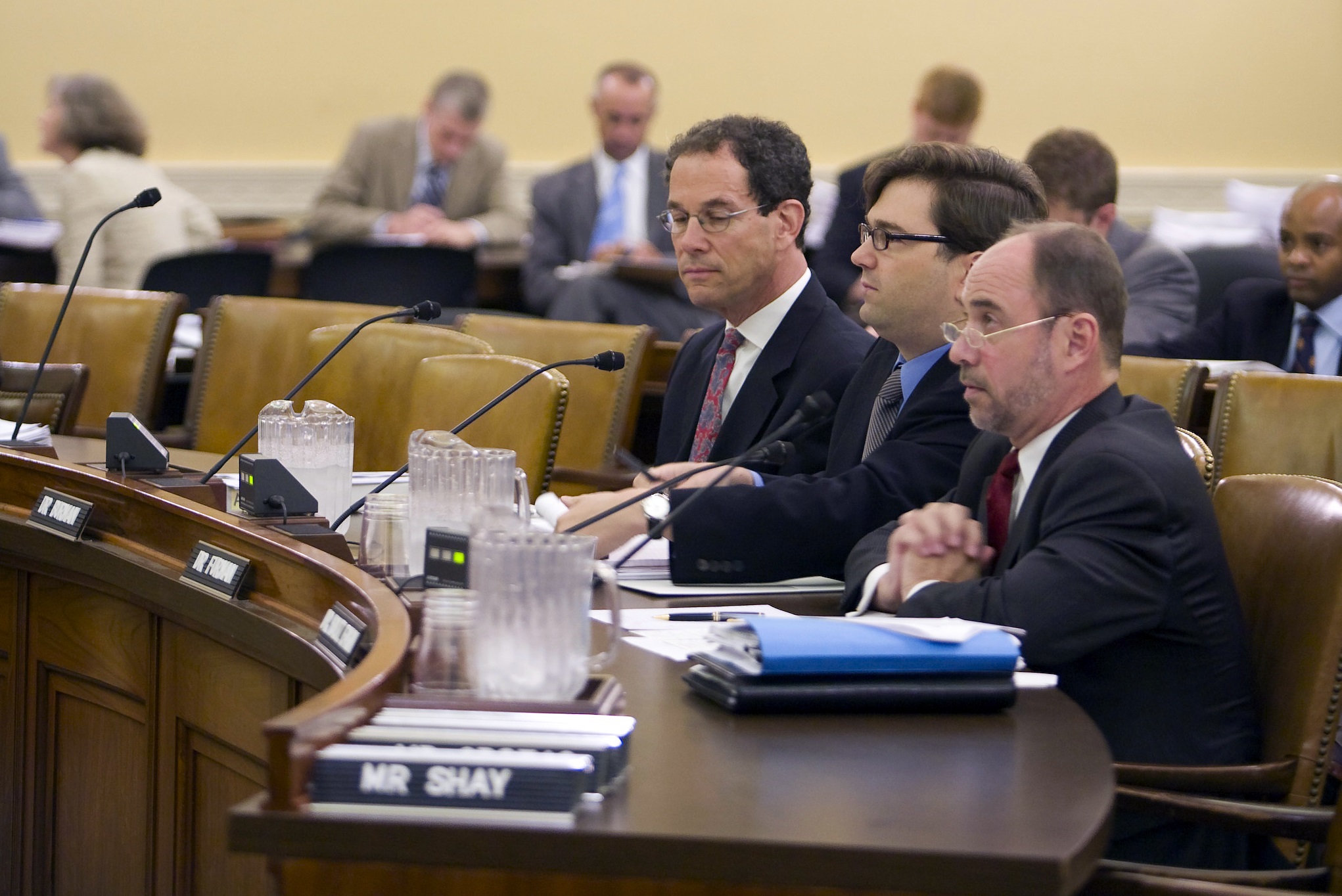 Three men sit in front of microphones in a room with additional people in the back.