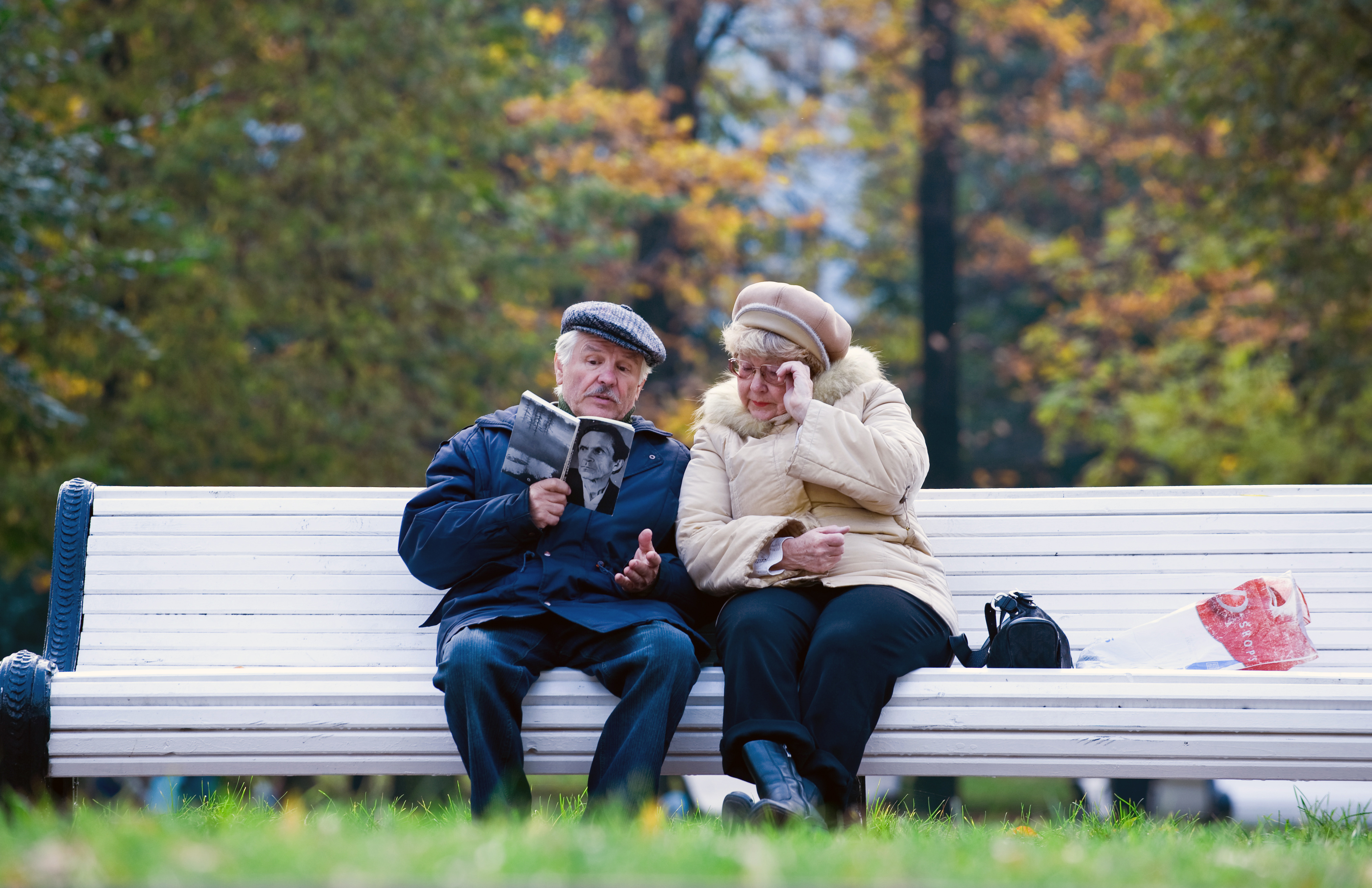 Elderly man and woman in coats read a book on a bench with trees in background