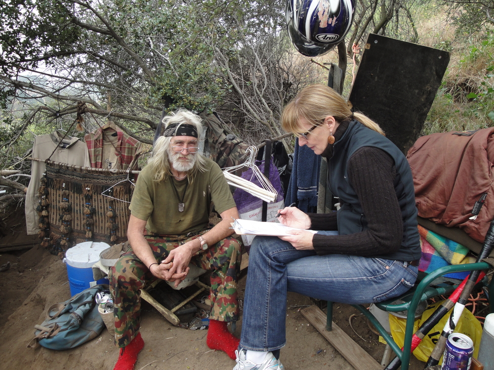 A man sits on a stool outside looking at the camera, next to a woman in a chair who is taking notes.