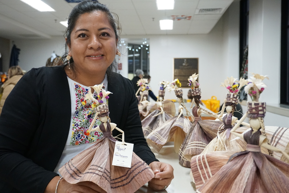 A woman holding a doll made from cornhusk smiles at the camera.