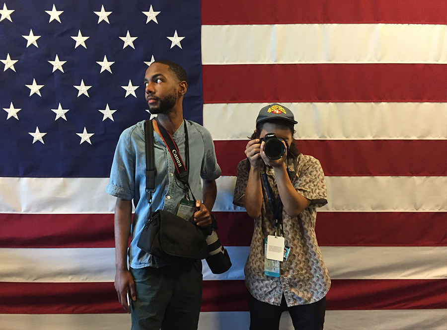Two people holding cameras and taking pictures in front of a flag.
