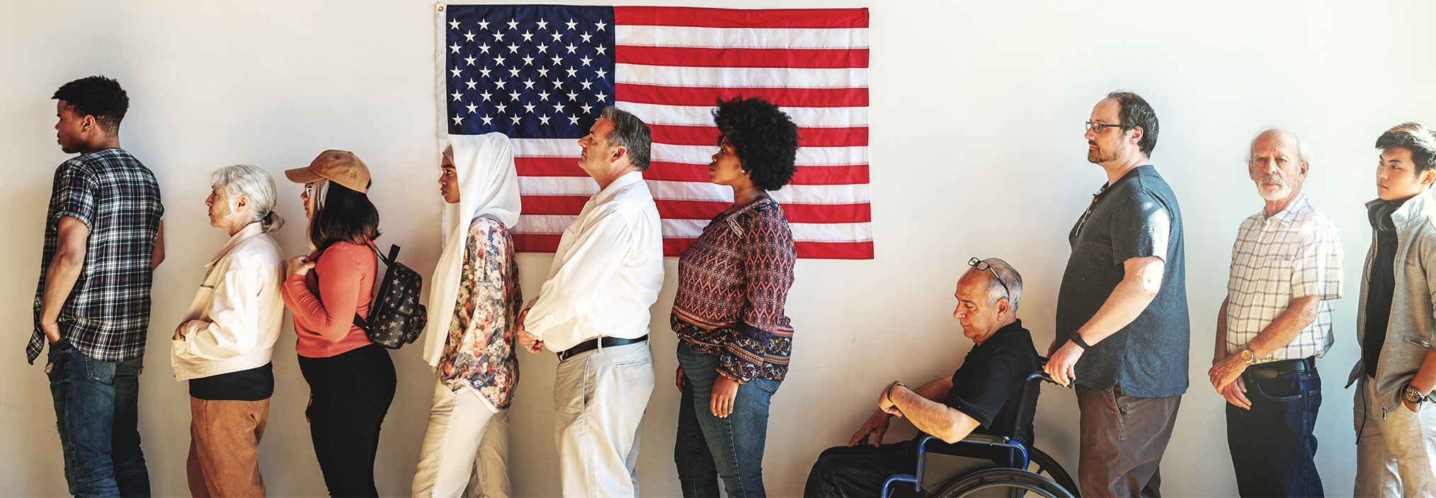 A diverse group of people line up in front of a US flag, including a person in a wheelchair.