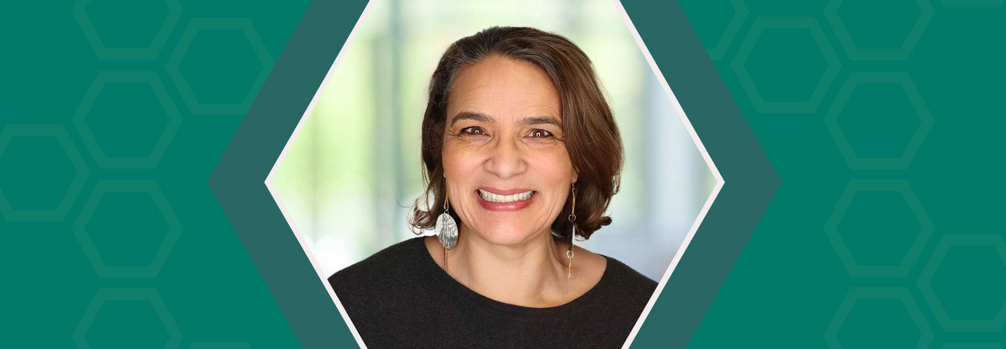 Smiling woman with brown hair in a black top, centered in a hexagon frame on a teal patterned background.