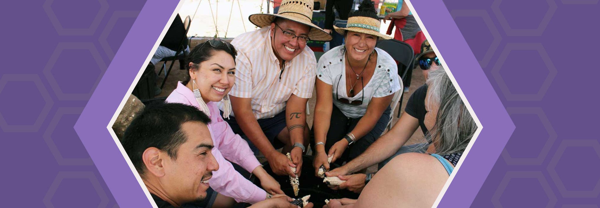 A group of six people seated in a circle holding corn cobs, smiling, and engaging in a communal activity.