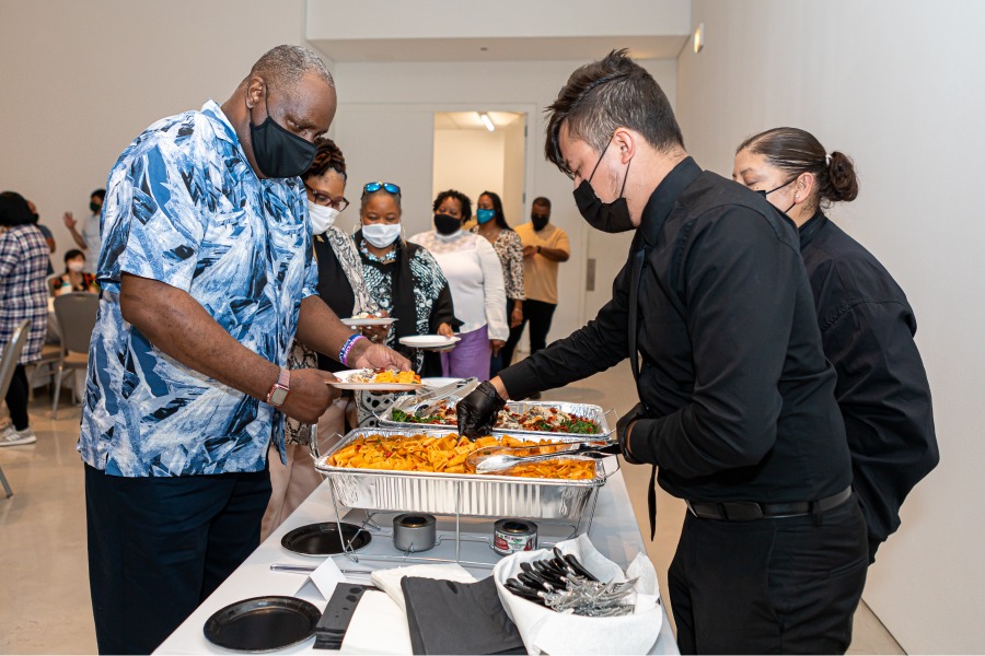 A man in a black shirt and pants serves another man in a blue shirt food at an event.