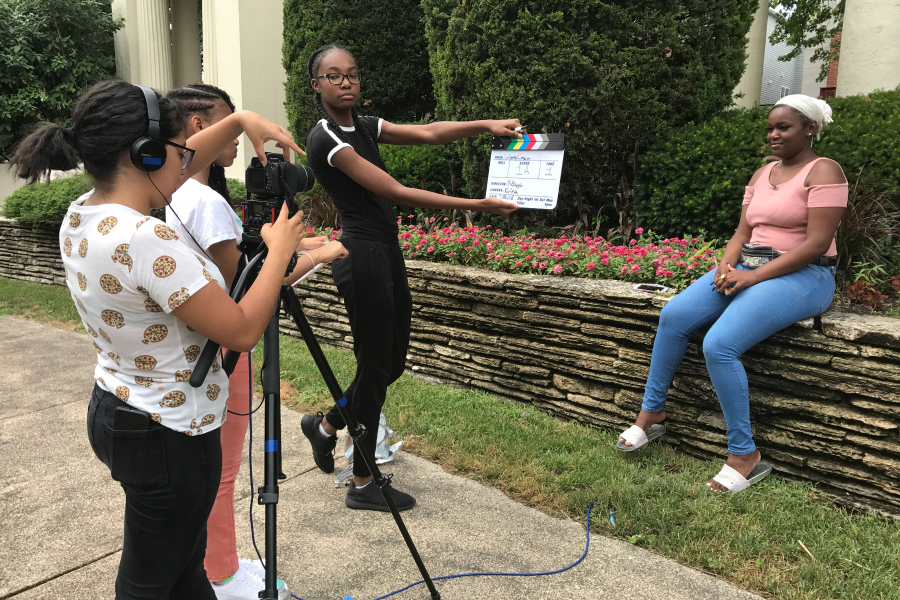 A girl is pointing a camera towards a girl sitting on a rock ledge while another girl holds up a clapboard in front of the camera.