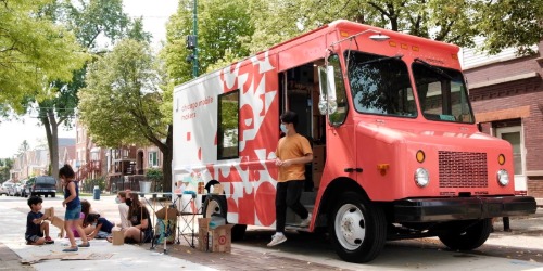 A red and white truck is parked on a street while people gather and sit on the sidewalk nearby under trees.