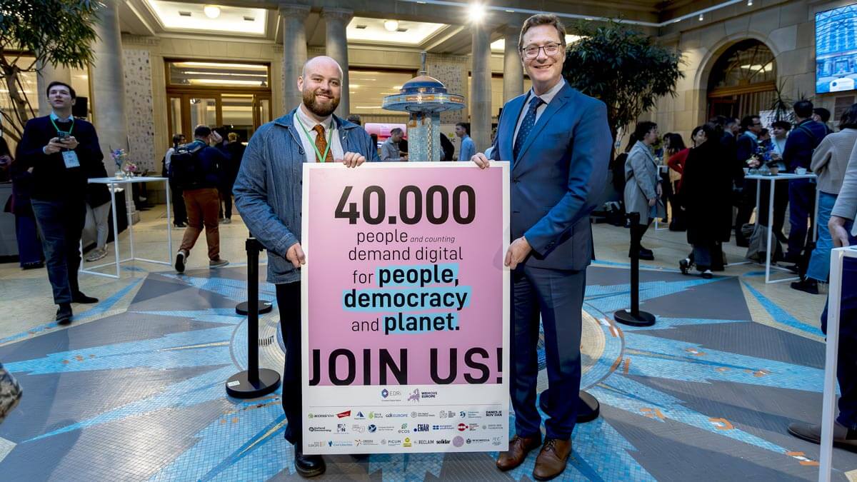 Two men hold a large sign with text that says, 40,000 people and counting demand digital for people, democracy and planet. JOIN US!, in a bright, open interior space.