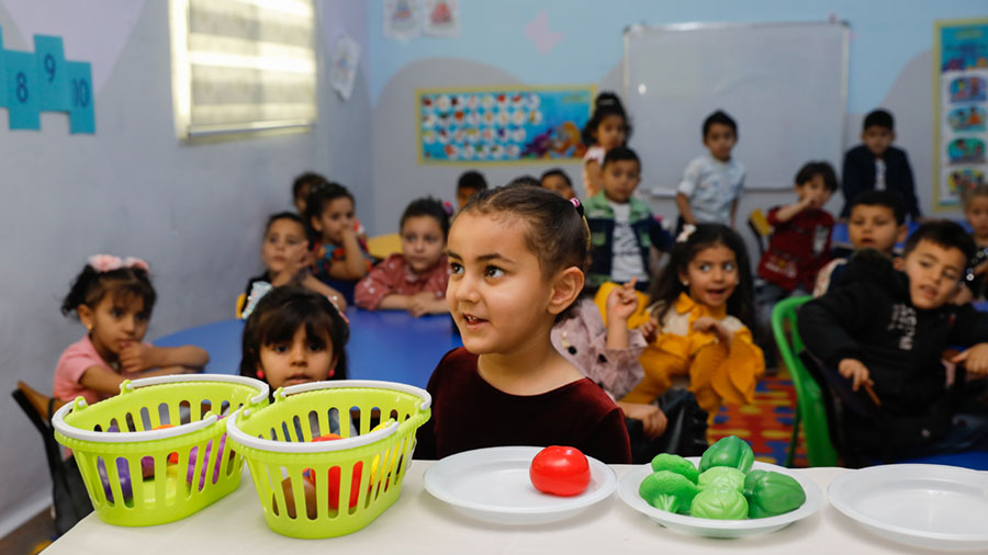 A young girl stands at a table with a group of children behind her.