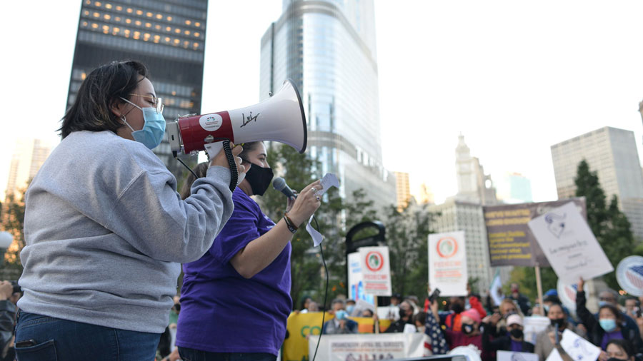 Two women wearing protective masks speaking to a crowd of people.