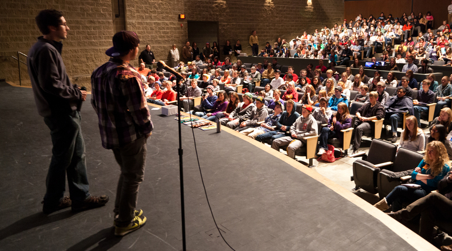 Two people stand on a stage in front of a large, seated crowd of people.