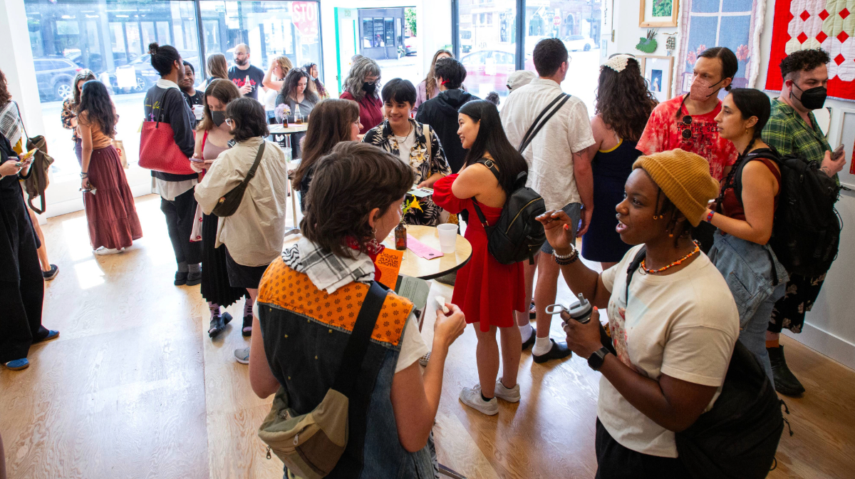 A diverse group of people socialize in a bright gallery space with art on the walls and refreshments on tables.