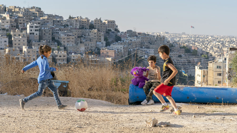 A young girl kicks a soccer ball to a young boy while another boy sits with a purple Muppet and watches.