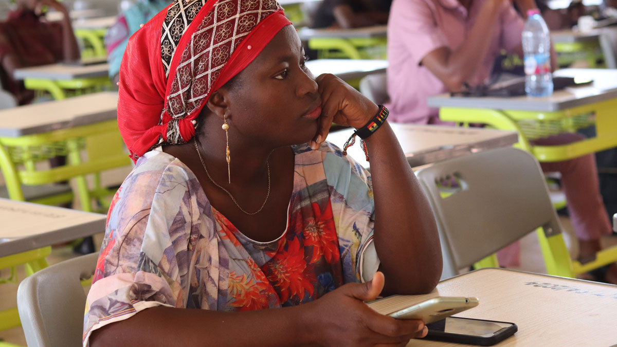 A young woman in a colorful blouse and red headscarf sits at a table, resting her chin on her hand while holding a smartphone, with other people in the background.