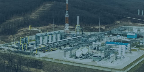 Aerial view of an industrial facility with machinery, tanks, a tall communications tower, and surrounding forested landscape.