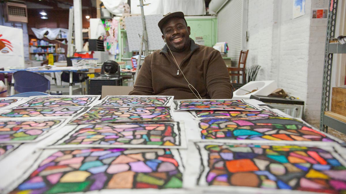 A man in a brown jacket and cap smiles at a table covered with colorful artworks in a studio.