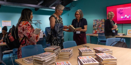 Four women stand and talk in a room with stacks of books on tables and a presentation screen in the background.