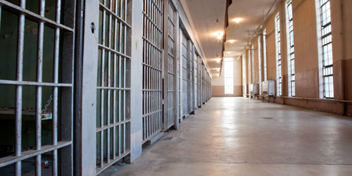 Prison corridor with barred cell doors and large windows.