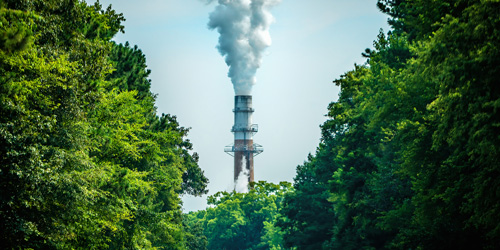 An electricity-generating facility smokestack releasing white smoke, surrounded by green trees under a blue sky.