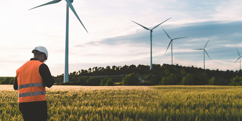 A person in a reflective vest stands in a field with wind turbines in the background.