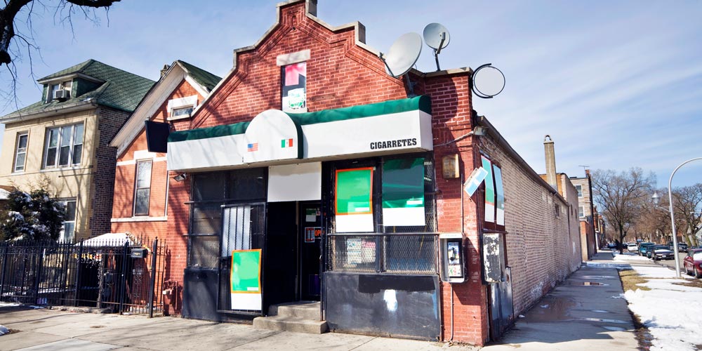 Red-brick corner shop with green-and-white awning, satellite dishes, neighboring houses, and snow along the sidewalk.