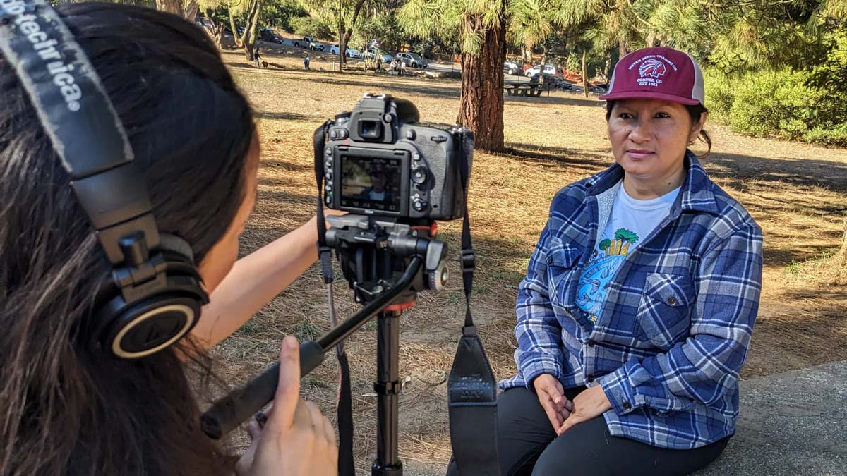 Person operating a camera records an interview with an individual seated in a park.