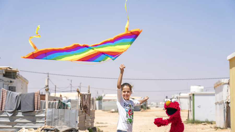 A girl flies a rainbow colored kite next to red furry muppet