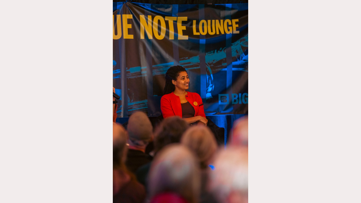 A close photo of a smiling woman, wearing a bright red cardigan. She sits during an interview at a music festival. 