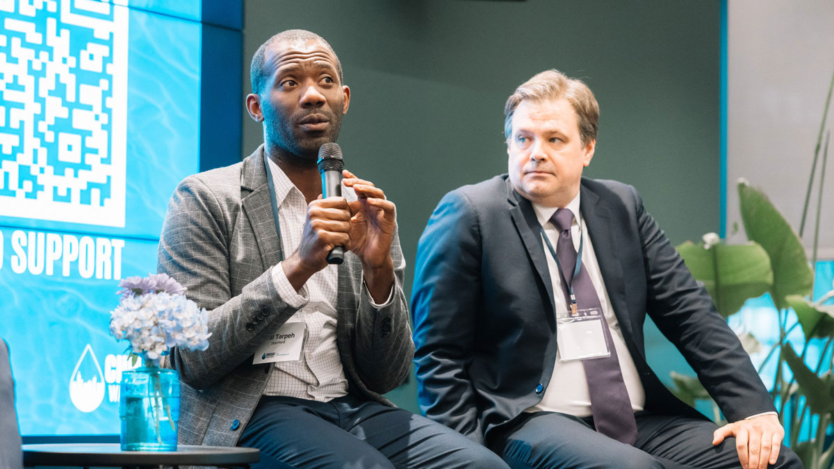 A close image of two men in suits at a public panel. The man on the left holds a microphone, mid-conversation while the man on the right listens. 