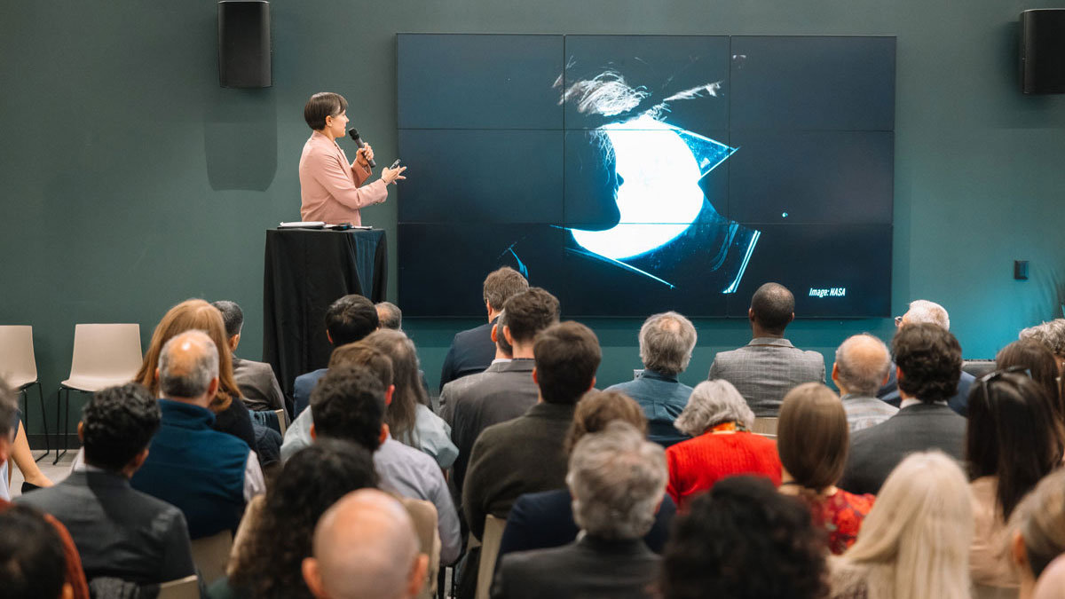 A crowd sits watching a video presentation as a speaker stands nearby, narrating. 