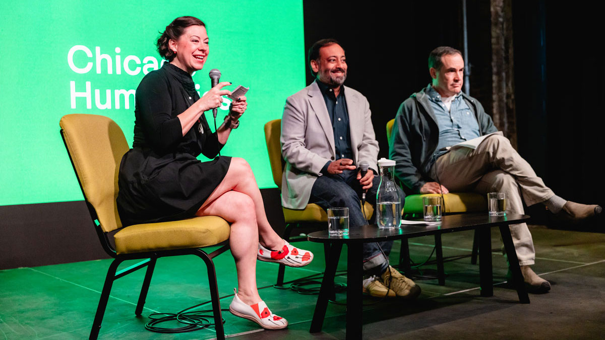 A smiling woman holds a microphone and notecards as she interviews two men. All three hold microphones and sit in front of a bright green screen that reads “Chicago Humanities.”