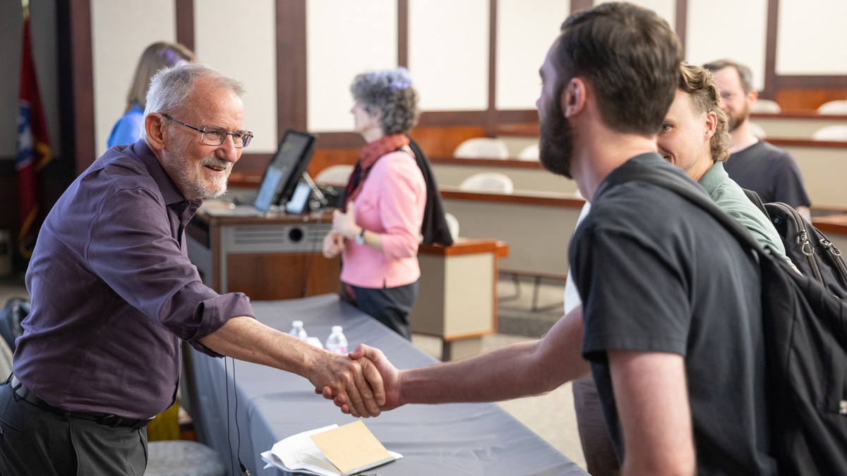 An older professor shakes hands with a younger college student. The two stand in a classroom, following a lecture.