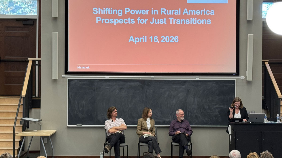 A trio of professors sits before an auditorium of students and community members. To the right of them, a moderator stands holding a microphone.