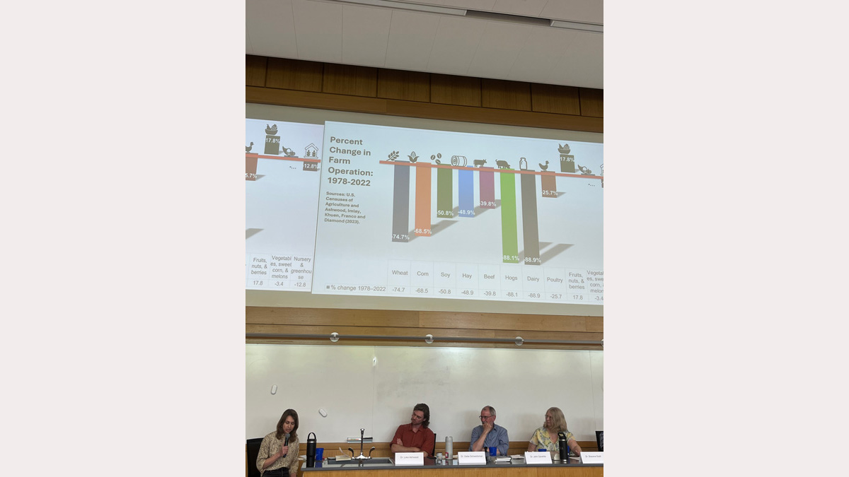 A woman reads from a notebook while three of her colleagues sit listening. Above the panel is a PowerPoint presentation displaying a colorful bar graph of information. 