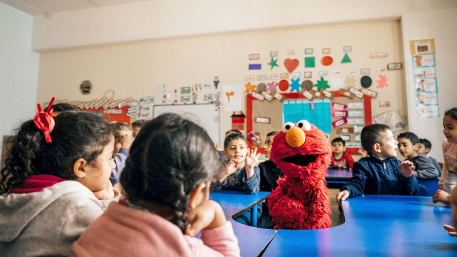 A group of kids sitting at a table in a classroom with Elmo the puppet