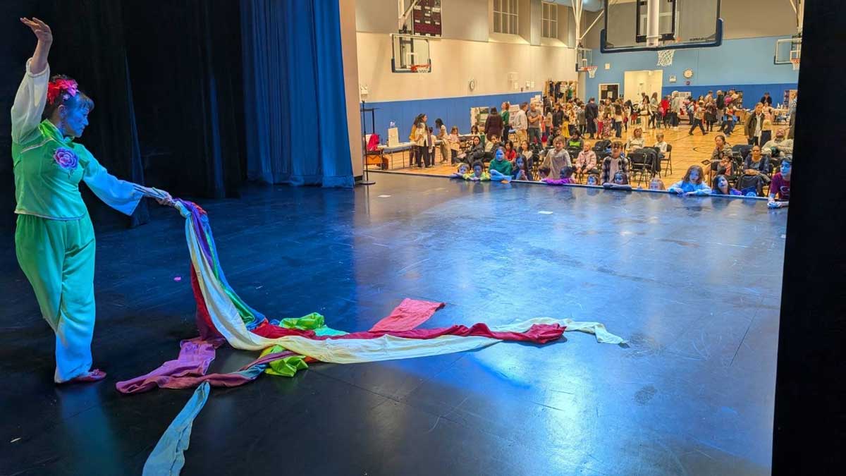 Performer on a stage with colorful fabric, facing an audience in a gymnasium.