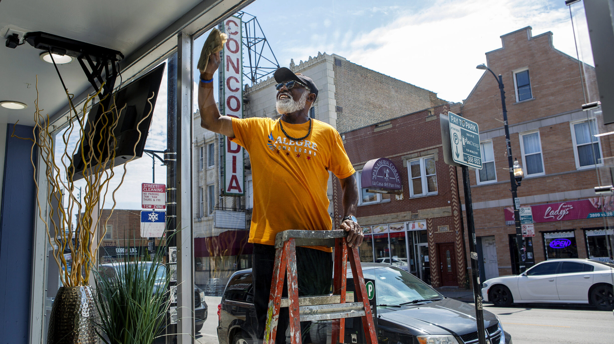 Man in a yellow shirt cleans a large window from a ladder on a sunny street with shops and cars.