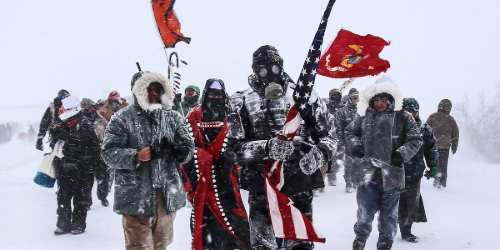 A group of people in winter clothing and masks march in heavy snow, carrying flags and banners.