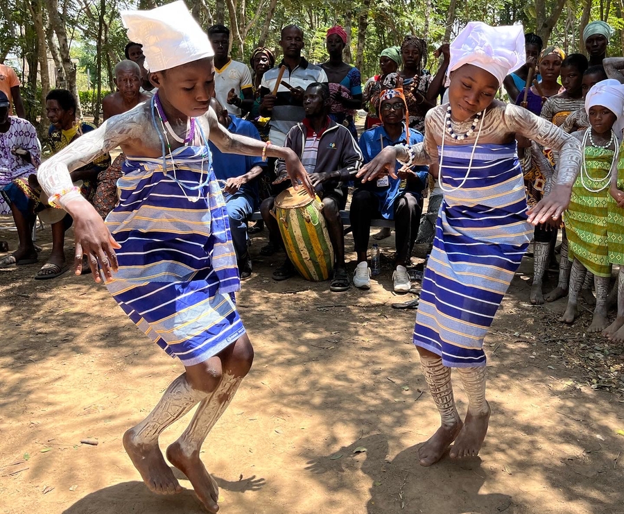 Two girls in blue and white dresses and white headpieces dance barefoot on dry ground