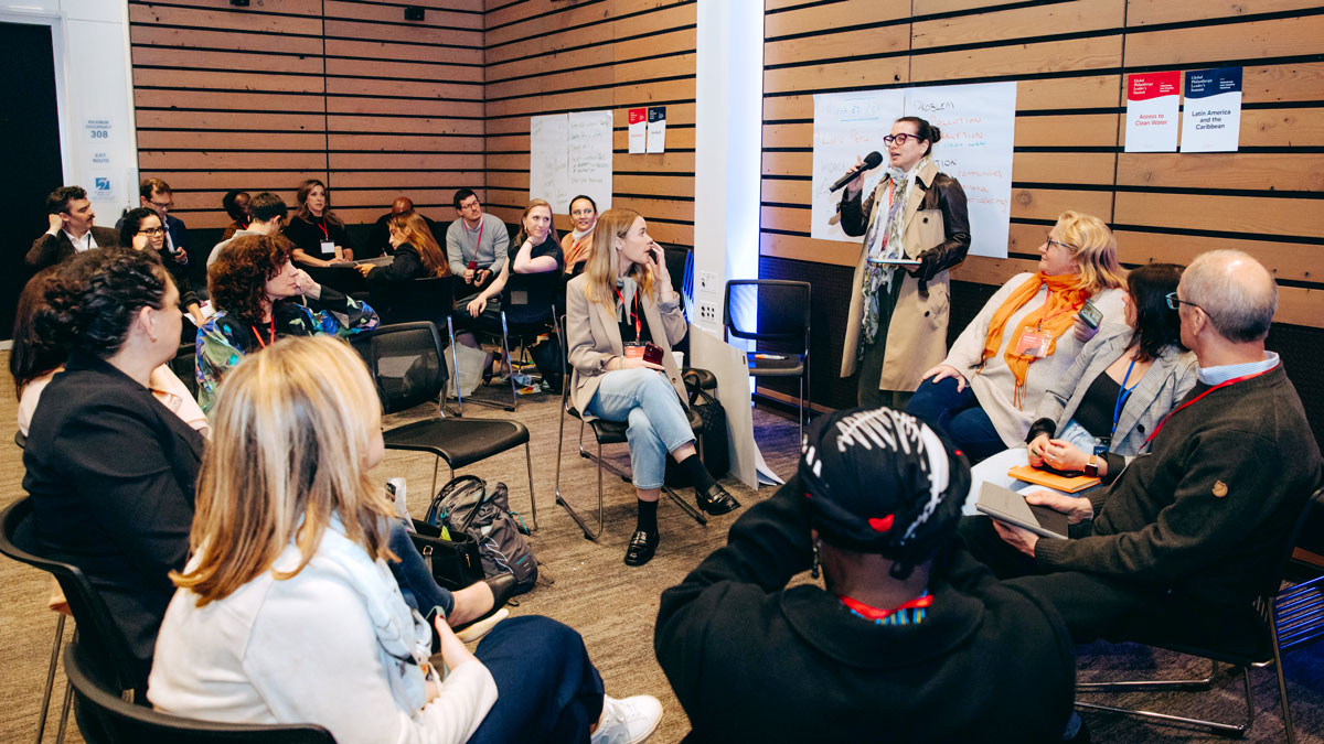 People in a meeting room with many raising their hands, and a speaker with a microphone standing.
