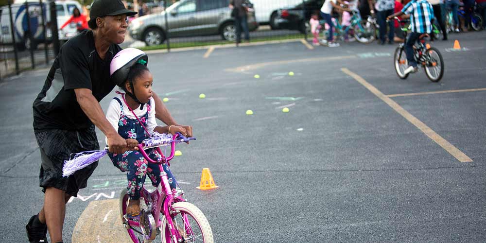 A father helps his young daughter balance on her pink bicycle. 
