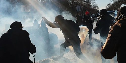 Protesters move through tear gas, with sunlight streaming through, creating a dramatic scene.