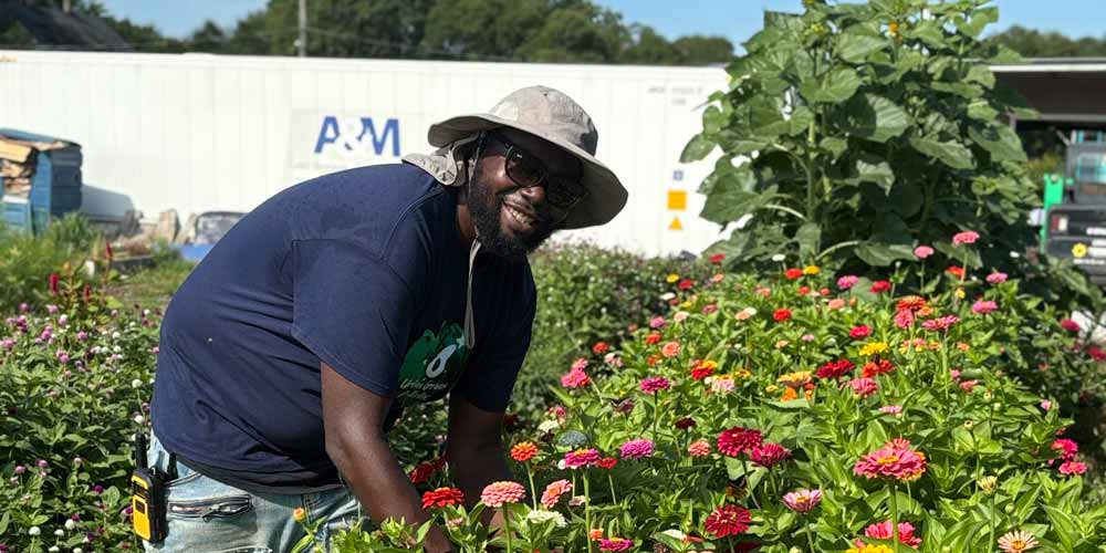 A man works in a community garden while smiling broadly. The man wears a navy blue shirt and wears a pale sunhat with a wide brim. The garden is full of orange blooms.