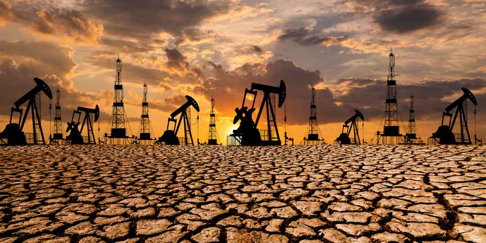 A view of an oilfield at sunset. In the background are the may silhouettes of oil drilling and refinery machines. In the foreground, the earth is dry and cracked as result of the drilling.